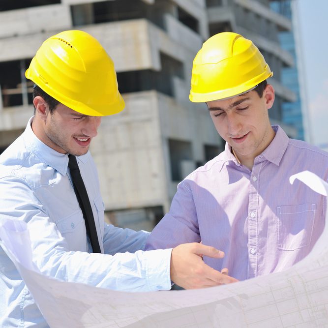 Two engineers wearing yellow helmets are discussing some plans